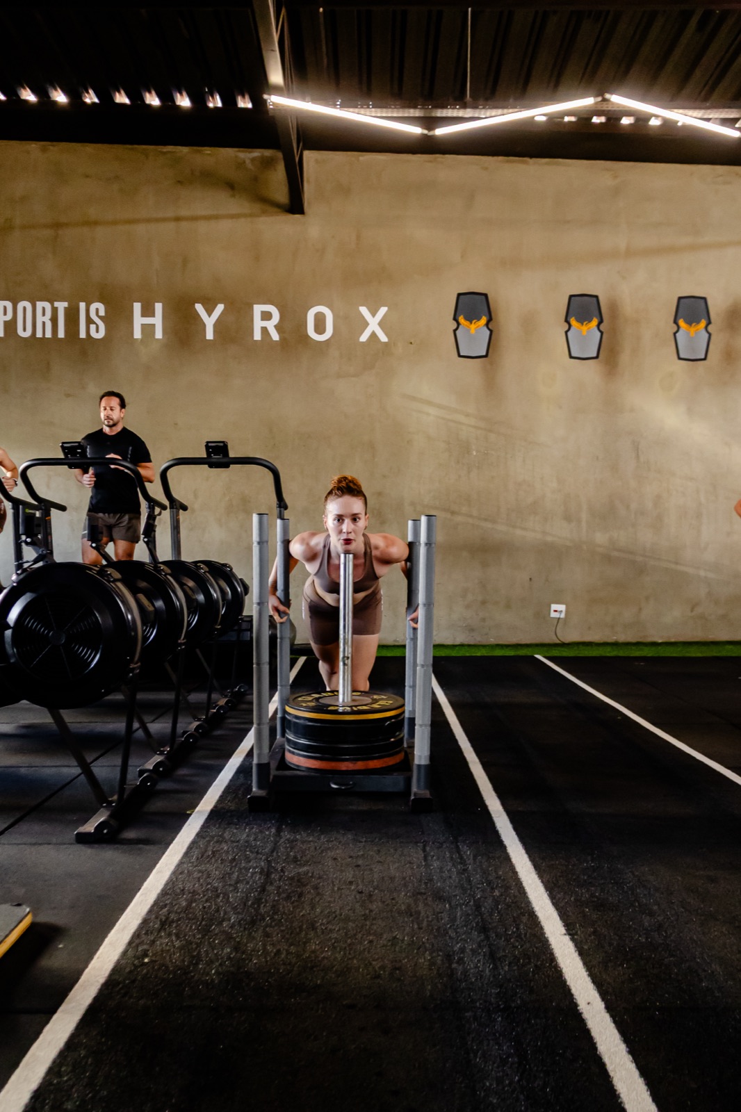 Aluna da Crane empurrando trenó de Hyrox, com a parede "Our sport is Hyrox" ao fundo
